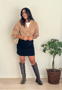 Beige kötött kardigán nagy gombokkal, fehér póló, fekete velúr mini szoknya és barna térdig érő csizma. Műnövény fonott kosárban a közelben.