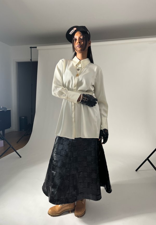 Camisa blanca de botones, falda negra texturizada, guantes negros con acentos de hardware, botas marrones y una gorra de béisbol negra.