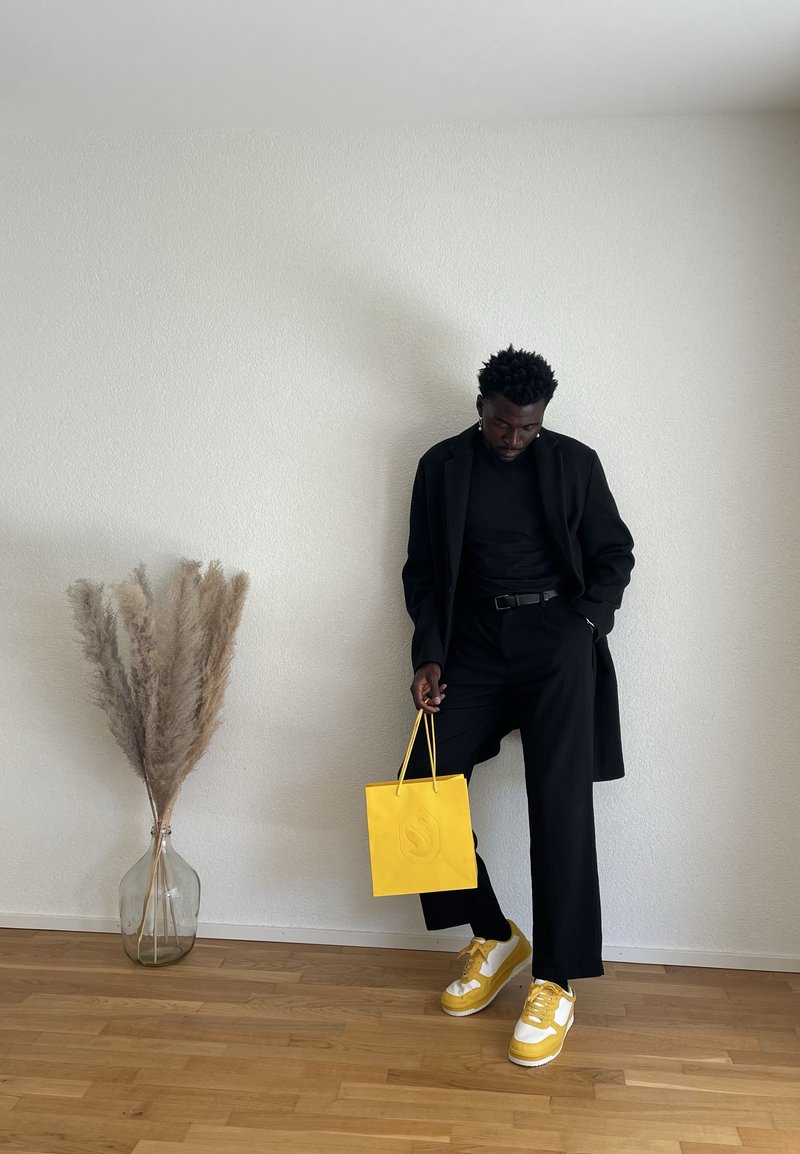 Homem em traje preto segurando uma bolsa de compras amarela, usando ténis amarelos combinando, ao lado de um vaso de vidro com erva-pampa.