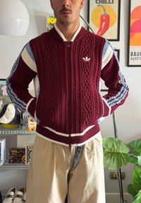 Tumepruun kootud jope lukuesiosaga, valgete ja hele siniste rõhutustega, mis sisaldab tekstuurset mustrit ja Adidas logo vasakul rinnal.