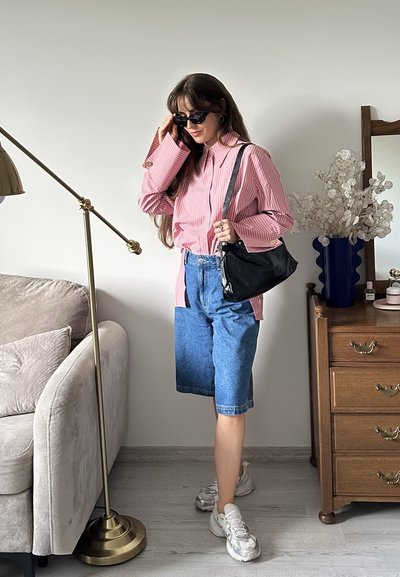 Camisa rosa a rayas, bermudas de mezclilla, bolso negro de hombro y zapatillas blancas. De pie junto a una lámpara y una mesa de noche de madera con decoración.