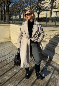 Beige trenchcoat med en svart pälsfäll, svarta högklackade stövlar, grå byxor och en svart handväska mot en utomhusbakgrund.