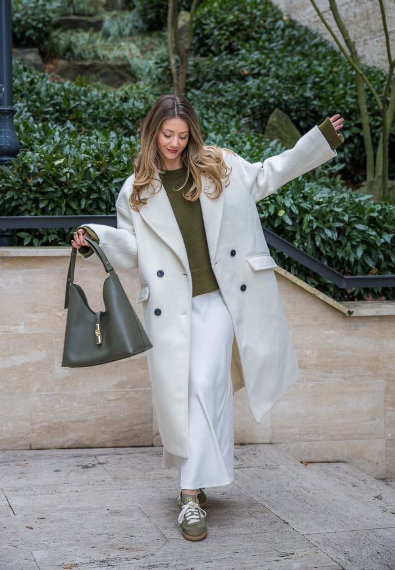 Femme en manteau blanc, pull olive et pantalon blanc tenant un sac à main olive, posant en plein air près de verdure et de marches en pierre.
