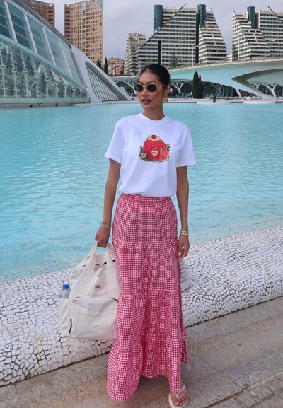 Camiseta blanca con un gráfico de postre de gelatina colorido, combinada con una falda larga de cuadros vichy roja en capas. Bolsa de tote beige y gafas de sol. Entorno al aire libre.