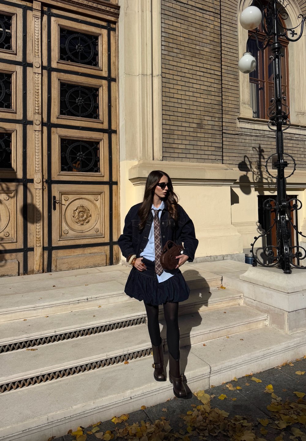 Giacca di jeans scura sopra una maglietta azzurra chiara e una gonna blu scuro con volant, abbinata a collant marroni e stivali al ginocchio. Borsa marrone e occhiali da sole.
