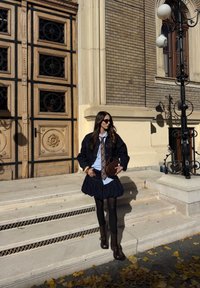 Giacca di jeans scura sopra una maglietta azzurra chiara e una gonna blu scuro con volant, abbinata a collant marroni e stivali al ginocchio. Borsa marrone e occhiali da sole.