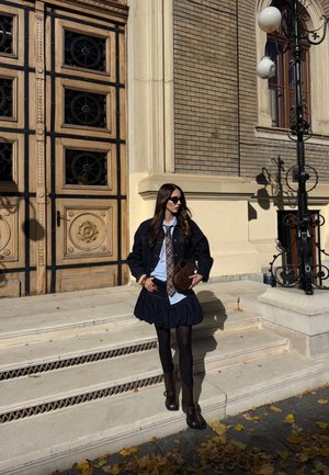 Dark denim jacket over a light blue shirt and a ruffled navy skirt, paired with brown tights and knee-high boots. Brown handbag and sunglasses.