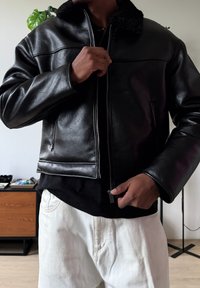 Veste en cuir noir avec col en fausse peau de mouton, coupe ajustée, fermeture éclair et poches frontales. Portée par-dessus une chemise noire et un pantalon blanc.
