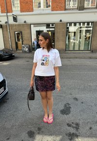 Mudel kannab valget T-särki, millel on värviline graafika, tumedat sädelevat mini-seelikut, roosasid plätusid ning hoiab käes väikest pruuni käekotti.