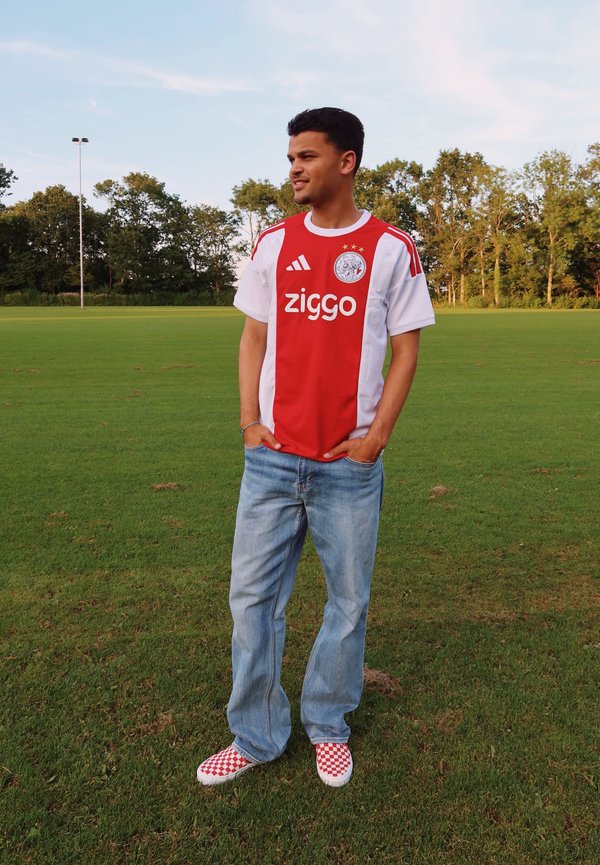 Giovane uomo in piedi su un campo d'erba che indossa la maglia da calcio dell'Ajax, jeans e scarpe slip-on rosse a quadri, guarda verso destra.
