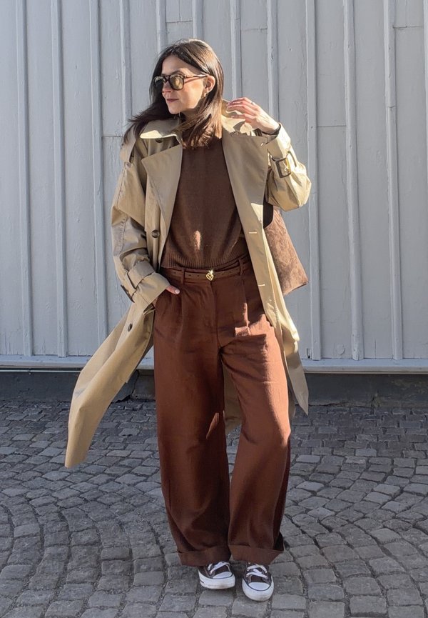 Γυναίκα με ανοιχτό μπεζ μπουφάν τύπου trench coat πάνω από καφέ πουλόβερ και φαρδιά καφέ παντελόνια, στέκεται σε λιθόστρωτο πεζοδρόμιο μπροστά σε λευκό τοίχο.