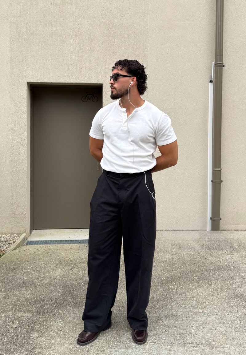 Homme portant des lunettes de soleil, un t-shirt blanc, un pantalon noir large et des écouteurs, debout les mains dans le dos près d'une porte grise avec un panneau vélo.