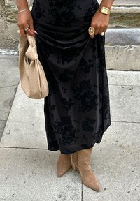 Robe noire à motif floral avec des détails froncés, associée à un sac à main en osier beige. Une main montre un bracelet en or et des bagues, avec des bottines beige.