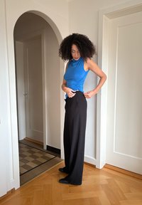 Haut bleu sans manches avec fronces sur le côté, associé à un pantalon noir taille haute. Sol en bois et à motifs visible en arrière-plan.