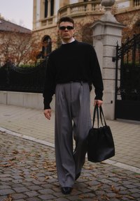 Černý svetr, šedé plisované kalhoty, černá kabelka. Hladká textura, moderní design. Sluneční brýle doplňují vzhled. Pozadí dlážděné ulice.