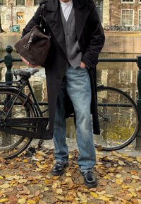 Svart trenchcoat över en grå cardigan, kombinerad med ljusblekta jeans och svarta läderskor. En brun väska hänger från axeln, i en parkmiljö.