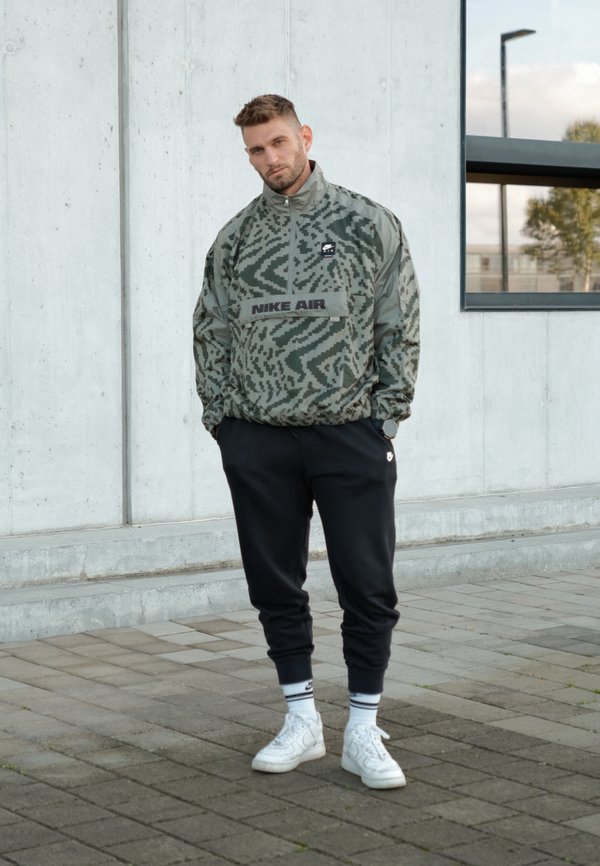 Chaqueta de camuflaje verde con un logo negro "NIKE AIR", combinada con pantalones deportivos negros y zapatillas blancas. Líneas limpias y ajuste atlético.