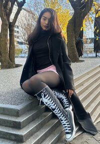 Schwarze kniehohe Schnürsneaker mit weißen Ösen, getragen mit schwarzen Strumpfhosen und einem rosa Rock, kombiniert mit einem langen schwarzen Mantel.