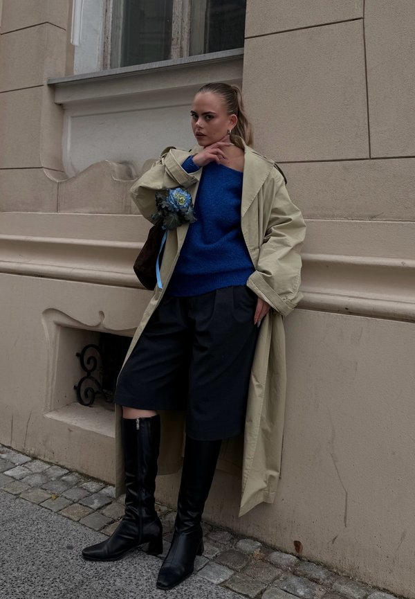 Une femme portant un trench-coat beige, un pull bleu, une jupe foncée et des bottes noires pose près d’un bâtiment en pierre, tenant un petit bouquet de fleurs bleues.