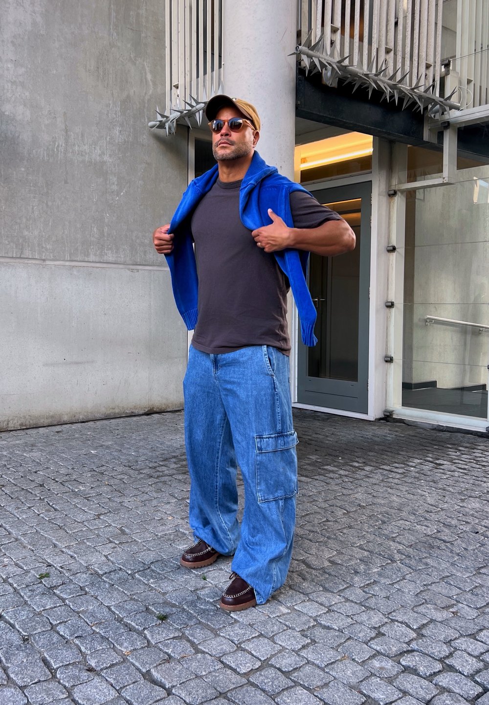 Felpa blu con zip posata sulle spalle, t-shirt grigia, pantaloni cargo in denim azzurri e larghi, scarpe marroni slip-on, occhiali da sole, in piedi all'aperto.