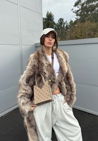 Femme portant un manteau en fourrure beige, un crop top blanc, un pantalon de survêtement gris, une casquette beige et tenant un sac à main beige à motifs en extérieur.