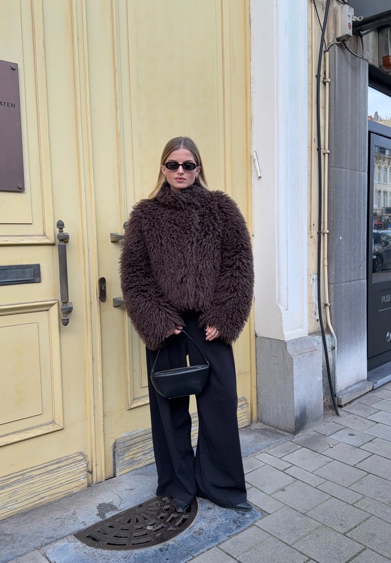 Manteau marron doux et ample avec col montant, pantalon noir à jambes larges, et petit sac à main noir ; accessoires minimalistes et lunettes de soleil.