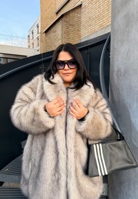 Femme portant de grandes lunettes de soleil noires et un manteau en fourrure beige, tenant un sac à main noir avec trois bandes blanches, debout sur des escaliers métalliques.