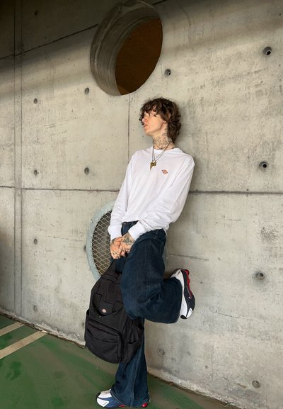 Camisa blanca de manga larga con un pequeño logo, jeans acampanados de mezclilla oscura, mochila negra y zapatillas blancas con acentos rojos. Fondo de pared de hormigón.