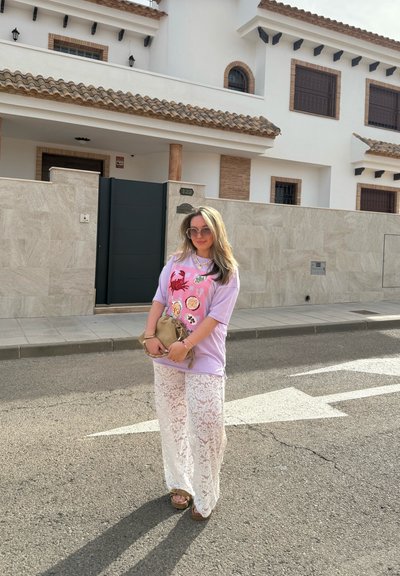 Camisa gráfica de color púrpura claro con diseños coloridos, combinada con pantalones anchos de encaje blancos. Sostenida una pequeña bolsa de mano beige. Calzado informal.