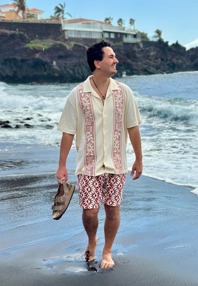 Hombre sonriendo y caminando descalzo en una playa de arena negra, sosteniendo sandalias, con olas del océano y casas en los acantilados al fondo.