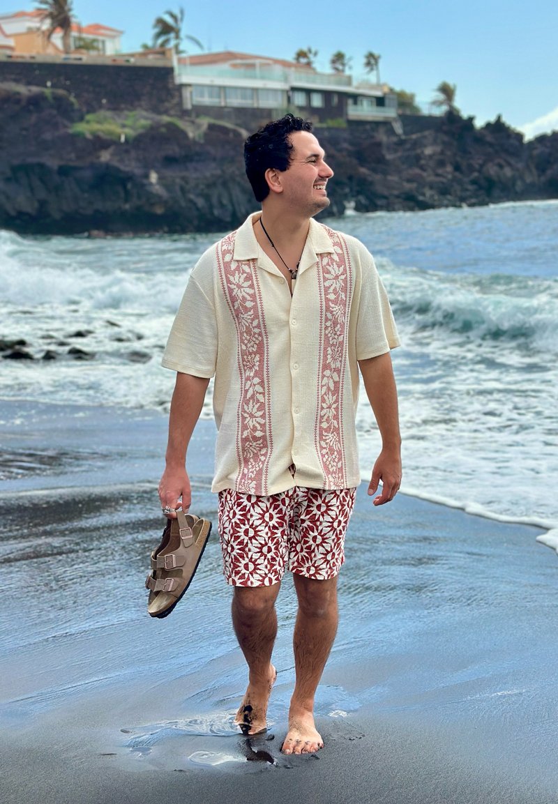 Uomo che sorride e cammina a piedi nudi su una spiaggia di sabbia nera, tenendo i sandali, con onde oceaniche e case sulla scogliera sullo sfondo.