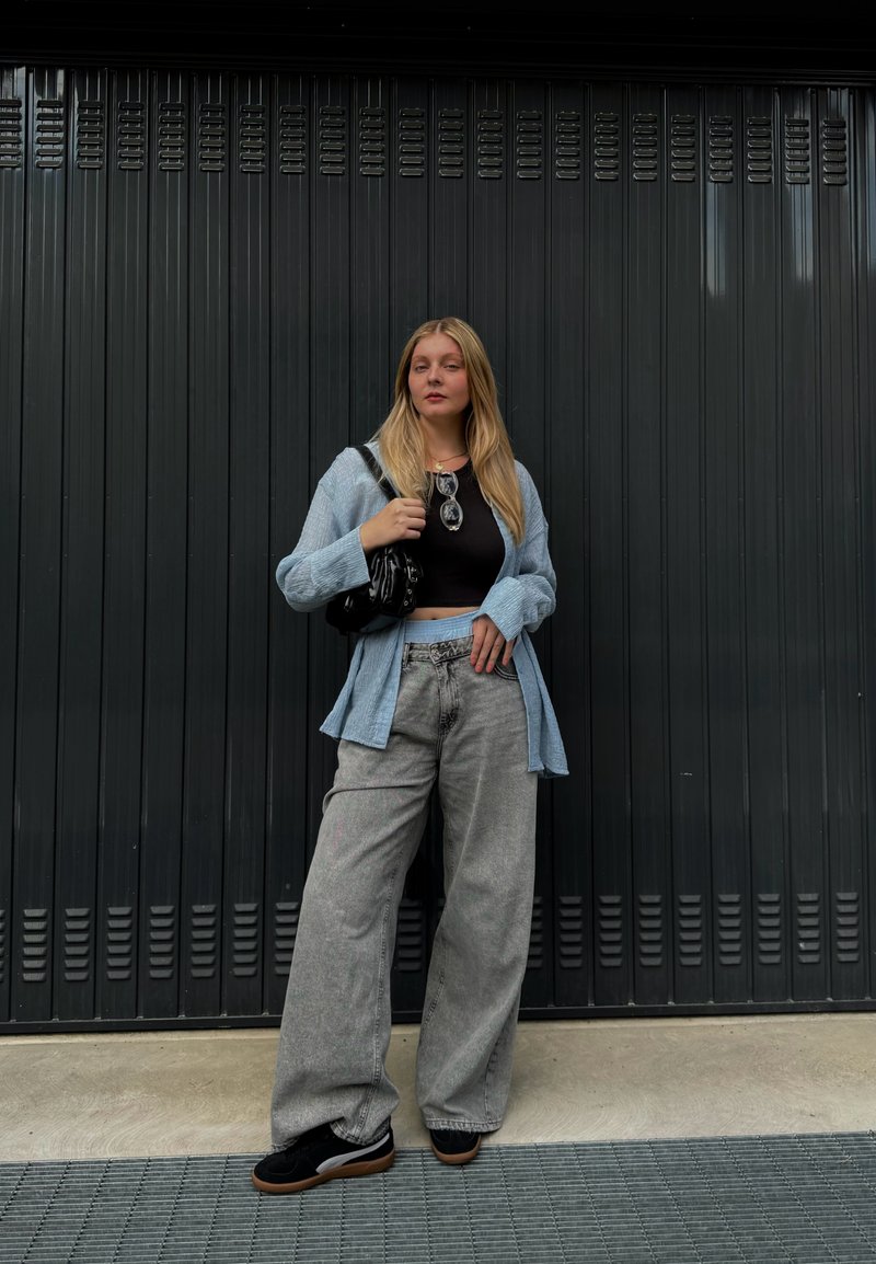 Ανοιχτό μπλε oversized πουκάμισο, μαύρο crop top, γκρι φαρδιά τζιν παντελόνια και μαύρα αθλητικά παπούτσια με σόλα από γόμα. Μαύρη τσάντα και γυαλιά ηλίου ως αξεσουάρ.