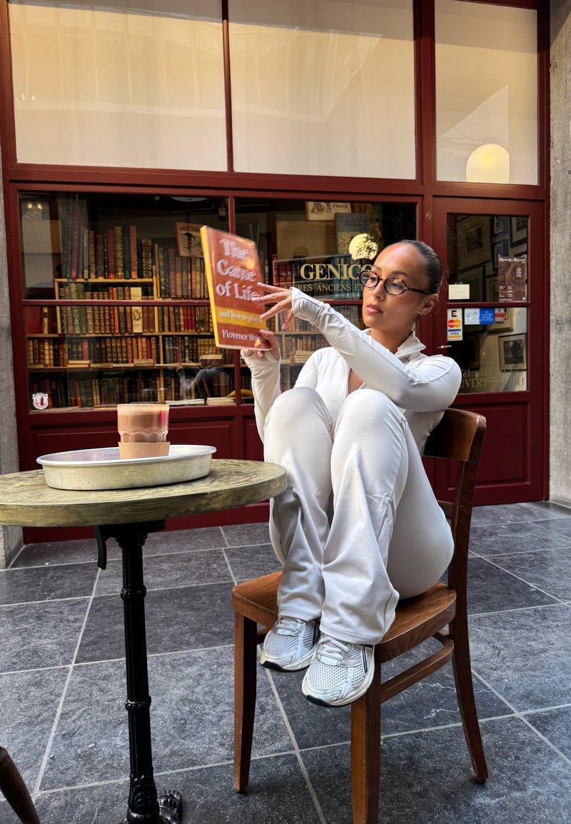 Femeie îmbrăcată în haine albe stând pe un scaun cu genunchii la piept, citind o carte, lângă o masă rotundă cu cafea, în fața feronierului unei librării.