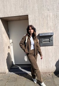 Tânără cu păr ondulat închis la culoare, purtând un costum maro din catifea și adidași albi, stă în picioare lângă un perete de piatră bej, aproape de o cutie poștală metalică.