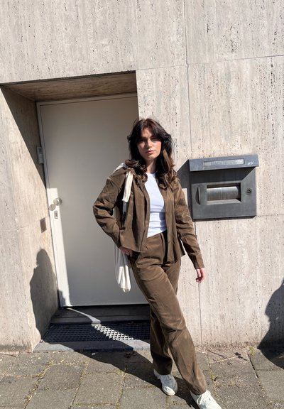 Mujer joven con cabello oscuro y ondulado, vistiendo un traje marrón de pana y zapatillas blancas, de pie contra una pared de piedra beige cerca de un buzón metálico.