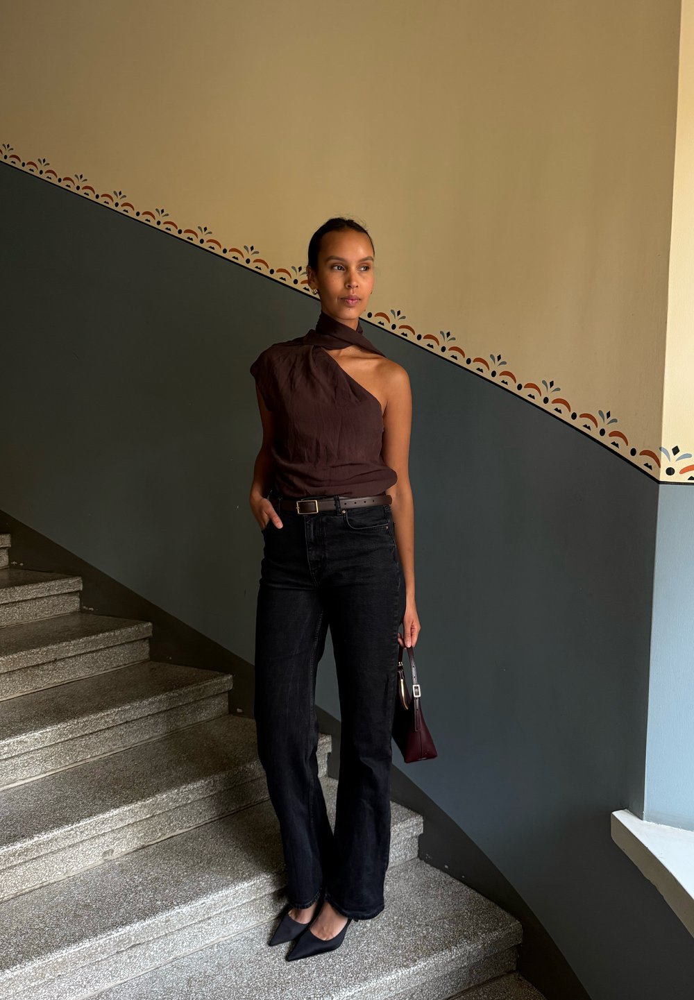 Haut asymétrique marron, jeans évasés noirs, talons noirs, tenant un sac bordeaux, se tenant sur des marches en pierre, devant un mur coloré.