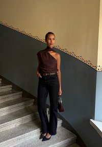 Haut asymétrique marron, jeans évasés noirs, talons noirs, tenant un sac bordeaux, se tenant sur des marches en pierre, devant un mur coloré.