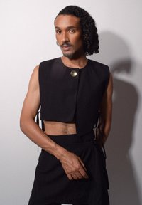 Svart kort topp med en rund guldknytnad. Har en strukturerad design och knyts på sidorna. Slät textur med minimalisk stil.