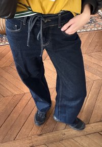 Pantalon en denim bleu foncé, coupe ample avec taille à cordon, coutures jaunes, poches, et texture lisse.