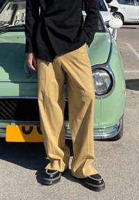 Calças largas beige combinadas com um blazer preto; sapatos pretos robustos com detalhes em corrente prateada. Carro vintage verde ao fundo.