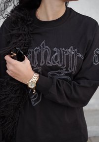 Melnā sporta jaka ar baltu šūtu tekstu, apvienota ar zelta pulksteni ar metāla siksniņu. Tur rokās melnu aksesuāru. Redzami teksturēti melni aksentu elementi.