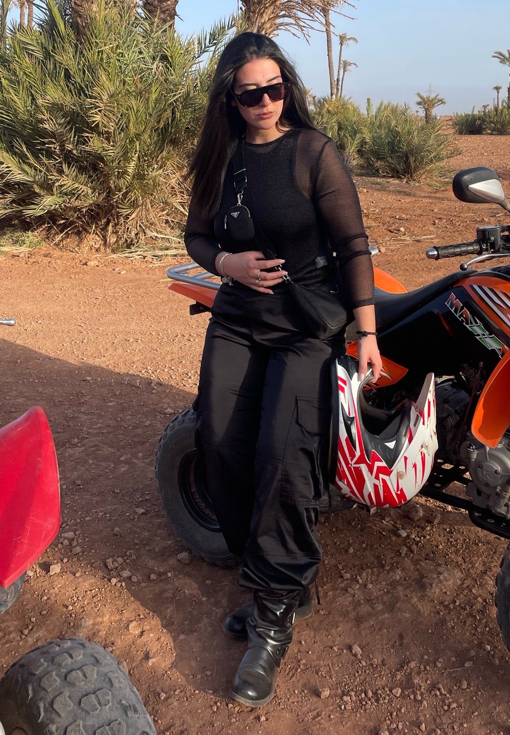 Maglietta nera trasparente a maniche lunghe, pantaloni cargo e stivali neri. Una piccola borsa nera e un casco rosso e bianco sono nelle vicinanze. Lo sfondo presenta palme.