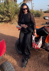 Maglietta nera trasparente a maniche lunghe, pantaloni cargo e stivali neri. Una piccola borsa nera e un casco rosso e bianco sono nelle vicinanze. Lo sfondo presenta palme.