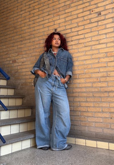 Chaqueta de denim con corte holgado, pantalones anchos de mezclilla de color azul claro, zapatos de patrón en blanco y negro, y collares superpuestos, frente a una pared de ladrillo.