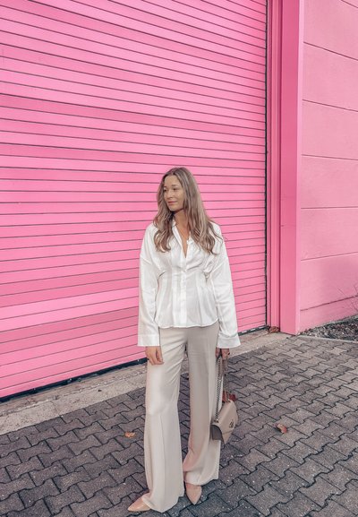 Camisa blanca de satén con botones y mangas largas, combinada con pantalones beige acampanados. Bolso beige y zapatos puntiagudos de color claro. Fondo rosa.