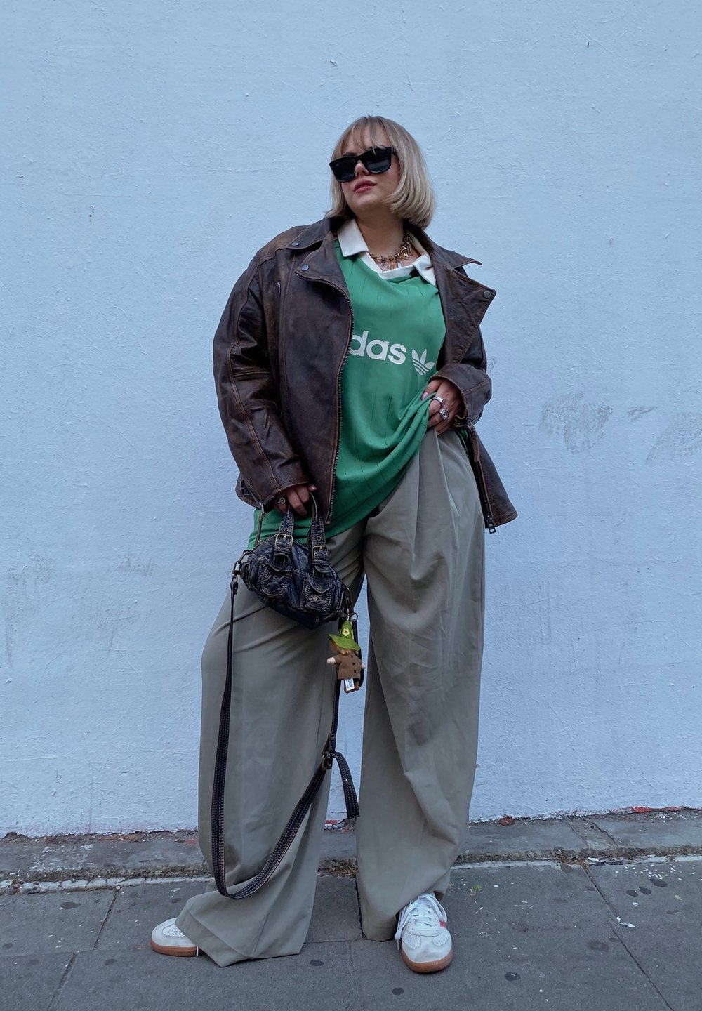 Καφέ δερμάτινο μπουφάν, πράσινη μπλούζα Adidas, υπερμεγέθεις ανοιχτό μπεζ παντελόνια, λευκά αθλητικά παπούτσια με καφέ σόλες, και ένα μοτίβο μαύρη τσάντα.