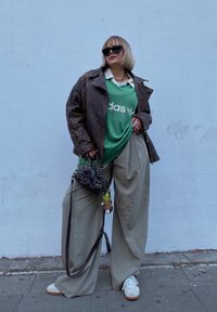 Καφέ δερμάτινο μπουφάν, πράσινη μπλούζα Adidas, υπερμεγέθεις ανοιχτό μπεζ παντελόνια, λευκά αθλητικά παπούτσια με καφέ σόλες, και ένα μοτίβο μαύρη τσάντα.