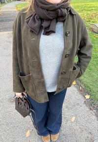 Semišová bunda olivově zelené barvy s velkými knoflíky a dvěma předními kapsami, nošená přes šedý svetr a zkombinovaná s tmavými džínovými kalhotami s rozšířenými nohavicemi a hnědými botami.