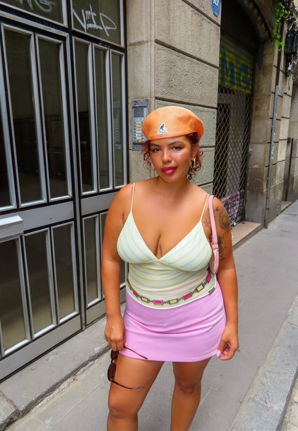 Femme portant un béret orange, un haut rayé jaune clair, une jupe rose et tenant des lunettes de soleil, debout sur un trottoir en ville près d'une entrée de bâtiment.