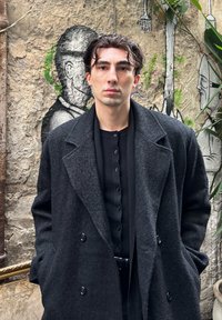 Lange zwarte jas met een gestructureerde afwerking, gedragen over een donker geribd shirt met knoopsluiting. Neutrale achtergrond met graffiti-elementen.